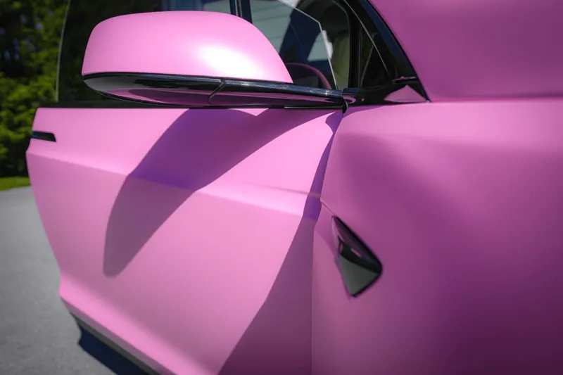 Close-up of a satin pink Tesla with the door slightly open, showing the wrapped mirror cap, the black flush door handle and film carried around the door edge