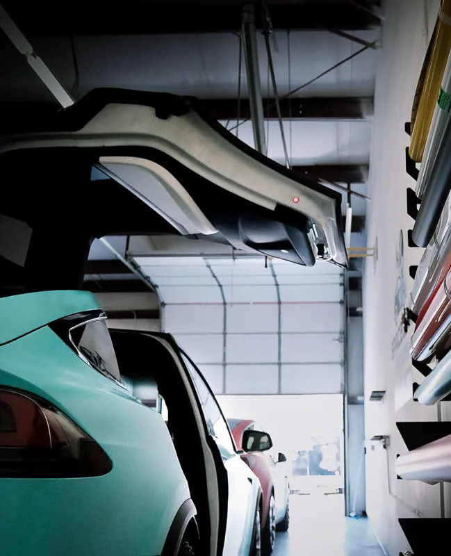 Rack of vinyl film rolls on the shop wall above a teal wrapped Tesla with its falcon-wing door raised